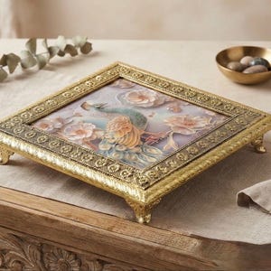 May include: A square decorative tray with an ornate gold-toned metal frame featuring intricate embossed patterns and four small feet. The centre panel displays a detailed illustration of a peacock surrounded by soft-toned floral blossoms.