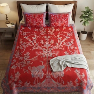 May include: A vibrant red bedspread and two matching rectangular pillow shams featuring a detailed print of white cranes, flowering branches, and birds. A folded light gray throw blanket rests on the lower right corner of the bedspread.