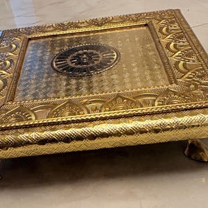 May include: A square gold-colored metal display stand featuring an intricate embossed border with repeating petal motifs and a central circular medallion. The platform has a textured surface and is supported by four small, curved metal feet.