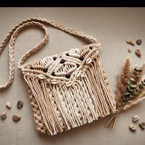 Puede incluir: Un bolso bandolera de macramé beige con correa trenzada, patrones de nudos complejos, cuentas de madera marrón oscuro y conchas de cauri blancas. La solapa frontal está decorada con largos flecos superpuestos en tonos tostados.