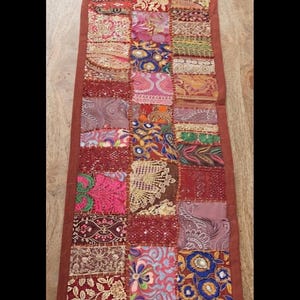May include: A rectangular table runner featuring a patchwork design of various fabric squares in shades of red, pink, gold, blue, and brown. The runner has a solid brown border and includes intricate embroidery, sequins, and floral patterns.