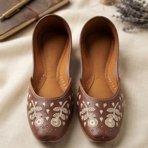 May include: A pair of brown slip-on flats featuring a rounded toe and a flat sole. The exterior of the shoes is decorated with intricate cream-colored floral embroidery patterns, including stems, leaves, and circular flower motifs.