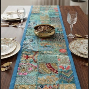 Può includere: Un runner da tavola rettangolare caratterizzato da un design patchwork composto da vari quadrati di tessuto blu, turchese e oro. Il runner è decorato con ricami complessi, motivi floreali e fili metallici, rifinito con un bordo blu.
