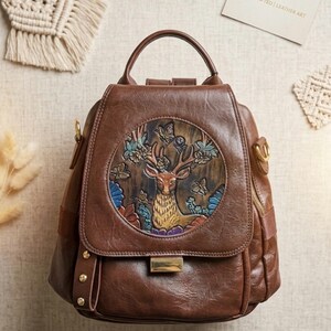 Puede incluir: Una mochila de cuero marrón con un panel circular en relieve en la solapa frontal. El panel muestra un ciervo rodeado de flores y mariposas en tonos azul, verde y morado. La bolsa tiene un asa superior, herrajes dorados y tachuelas laterales.