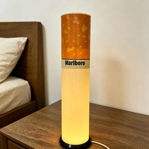 Può includere: Una lampada da tavolo cilindrica progettata per assomigliare a una sigaretta. La sezione superiore presenta un motivo a filtro arancione maculato, mentre quella inferiore emette una luce calda. Una fascia bianca riporta la scritta Marlboro.