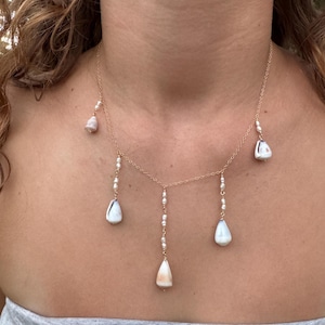 Puede incluir: Un collar de cadena en tono dorado con cinco dijes colgantes, cada uno compuesto por una pequeña concha marina blanca suspendida de una corta hilera de diminutas perlas de semillas blancas. El collar tiene un diseño de eslabones finos.