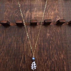 Peut inclure: Un collier à fine chaîne dorée avec un pendentif composé d'une perle gris foncé placée au-dessus d'un petit coquillage blanc orné d'un motif à grille noire. Le collier pend verticalement, mettant en valeur le contraste entre la chaîne dorée, la perle lisse et le coquillage texturé.