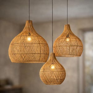 May include: A set of three hanging pendant lights featuring woven tan rattan shades in a teardrop shape. Each shade has an open lattice pattern and is suspended by a thin black cord, with a single glowing light bulb visible inside each fixture.
