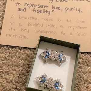 Puede incluir: Dos clips para zapatos plateados con pedrer&iacute;a azul y transparente en una peque&ntilde;a caja verde. La caja tiene una nota escrita a mano que dice "para representar el amor, la pureza y la fidelidad" y "Una hermosa pieza para el zapato de una hermosa novia, para representar los componentes clave para un matrimonio s&oacute;lido."