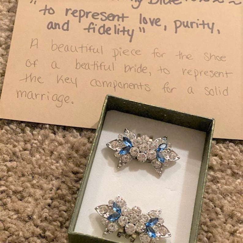 Puede incluir: Dos clips para zapatos plateados con pedrer&iacute;a azul y transparente en una peque&ntilde;a caja verde. La caja tiene una nota escrita a mano que dice "para representar el amor, la pureza y la fidelidad" y "Una hermosa pieza para el zapato de una hermosa novia, para representar los componentes clave para un matrimonio s&oacute;lido."