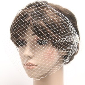 May include: A white, diamond-patterned birdcage veil, a bridal accessory, is draped over a mannequin's head. The veil is secured with small, pearl-like embellishments. The mannequin has brown hair and a neutral expression.