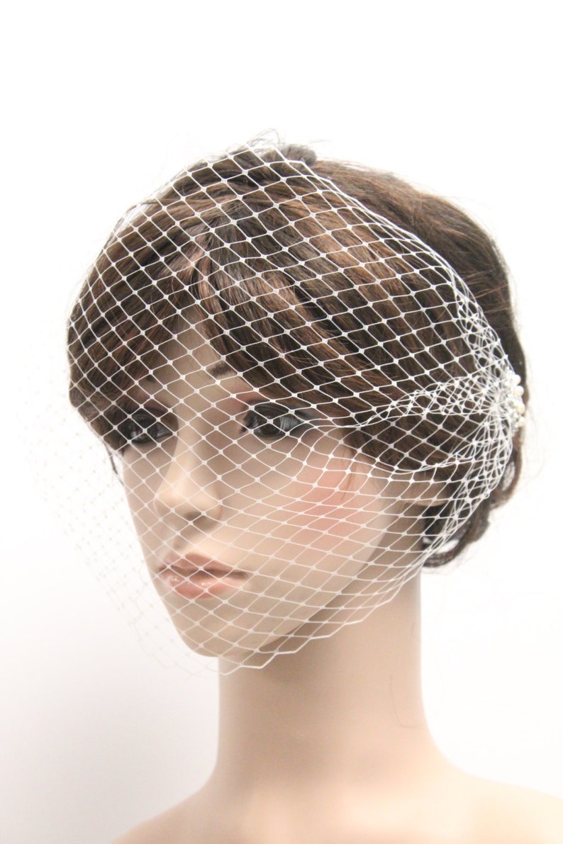May include: A white, diamond-patterned birdcage veil, a bridal accessory, is draped over a mannequin's head. The veil is secured with small, pearl-like embellishments. The mannequin has brown hair and a neutral expression.