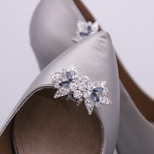 Puede incluir: Un par de zapatos de tac&oacute;n alto de sat&eacute;n plateado con un adorno de joyas en la punta. El adorno es una flor plateada con diamantes de imitaci&oacute;n transparentes y azules.