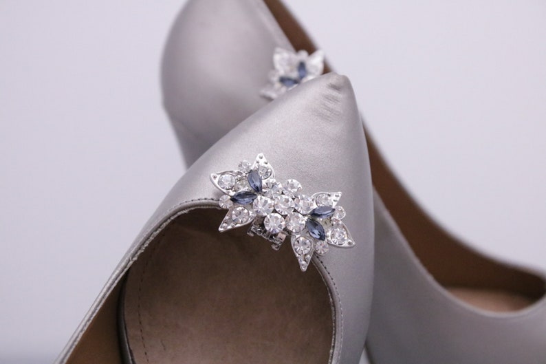 Puede incluir: Un par de zapatos de tac&oacute;n alto de sat&eacute;n plateado con un adorno de joyas en la punta. El adorno es una flor plateada con diamantes de imitaci&oacute;n transparentes y azules.