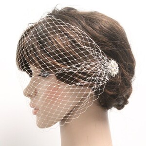 May include: A white birdcage veil with a diamond-shaped net pattern. The veil is attached to a hair clip adorned with small, round, pearl-like beads. The hair accessory is positioned on the side of the head, near the ear.