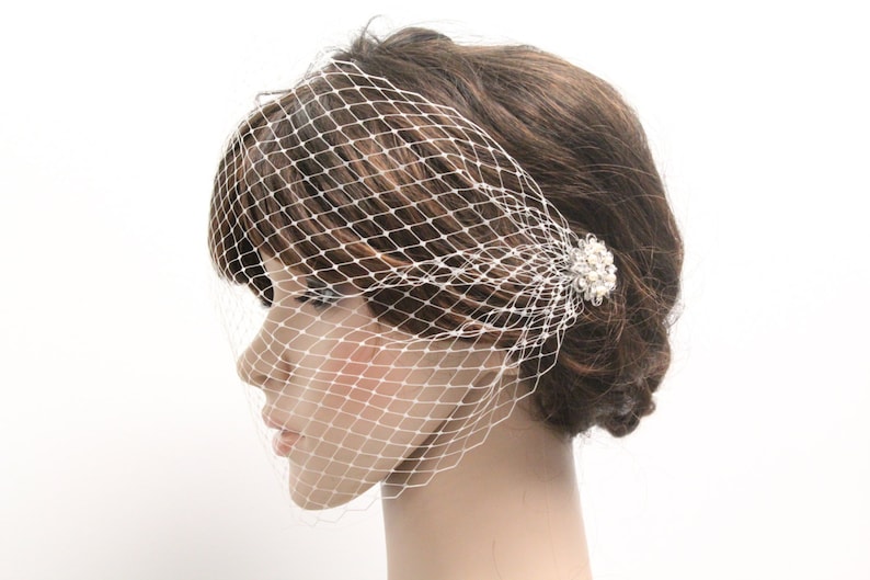 May include: A white birdcage veil with a diamond-shaped net pattern. The veil is attached to a hair clip adorned with small, round, pearl-like beads. The hair accessory is positioned on the side of the head, near the ear.