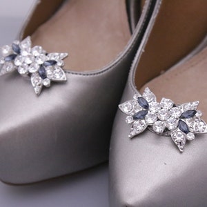 Puede incluir: Un par de zapatos plateados con un adorno de cristal y strass azul oscuro en la punta.