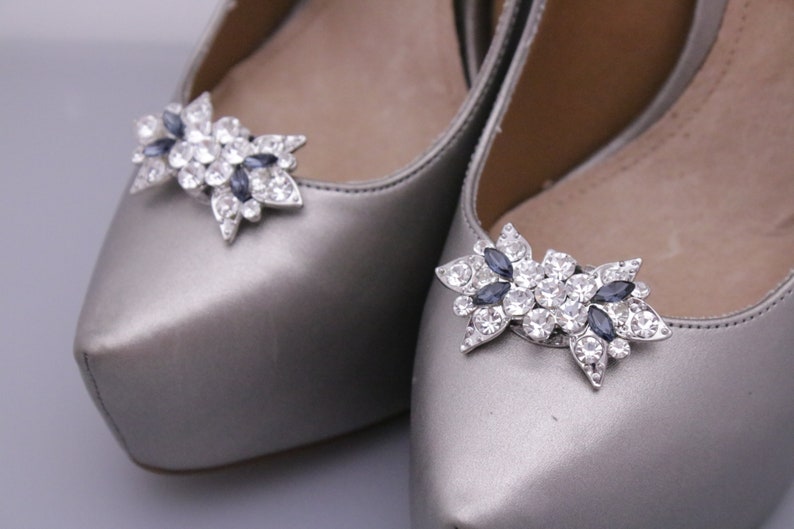 Puede incluir: Un par de zapatos plateados con un adorno de cristal y strass azul oscuro en la punta.