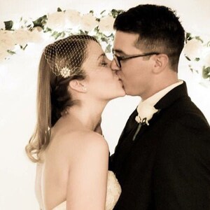 May include: A couple shares a kiss during a wedding ceremony. The bride wears a veil and a strapless dress. The groom is in a black suit with a white flower on his lapel. A floral arch is in the background.