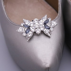 Puede incluir: Un par de zapatos de sat&eacute;n plateado con un adorno de joyas en la punta. El adorno es un lazo plateado con diamantes de imitaci&oacute;n transparentes y azul oscuro.