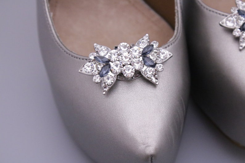 Puede incluir: Un par de zapatos de sat&eacute;n plateado con un adorno de joyas en la punta. El adorno es un lazo plateado con diamantes de imitaci&oacute;n transparentes y azul oscuro.