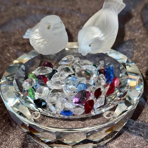 May include: A clear crystal bowl with a faceted exterior edge, containing a collection of small, multi-colored faceted glass gems in shades of red, blue, green, purple, and clear. Two frosted glass bird figurines are perched on the bowl rim.