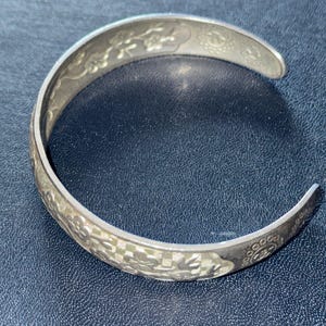 May include: A silver-toned metal cuff bracelet featuring an embossed pattern of cherry blossoms and branches. The surface displays a textured checkered background behind the floral motifs, with circular decorative stamps at the open ends of the band.