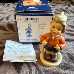 Könnte beinhalten: Eine Goebel Hummel Porzellanfigur mit dem Titel First Mate #1850, die ein Kind mit Matrosenmütze, orangefarbenem Hemd und blauer Hose zeigt, das ein Ruder hält. Der Artikel enthält die originale blaue Pappschachtel mit EAN 4005169018503 und ein Zertifikat.