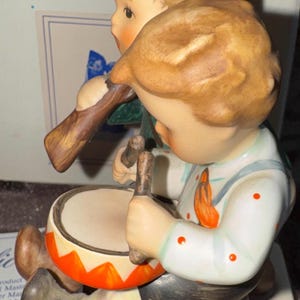 May include: A ceramic figurine featuring two children, one wearing a red hat and playing a wooden flute, and the other wearing a white shirt with orange polka dots and playing a drum with orange and white triangular patterns on the side.