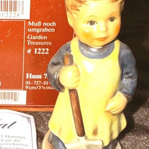 Könnte beinhalten: Eine Keramikfigur eines Kindes mit blonden Haaren, das eine gelbe Schürze über einem dunkelblauen Hemd trägt und eine Schaufel hält. Die Figur steht auf einem runden Sockel vor einer Schachtel mit der Aufschrift Muß noch umgraben Garden Treasures # 1222.