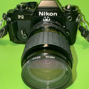May include: A black Nikon FG 35mm film camera featuring a textured grip, a mounted zoom lens with a rubberized focus ring, and a 52mm Hoya filter attached to the front. The camera body includes silver metal strap lugs and various manual dials.