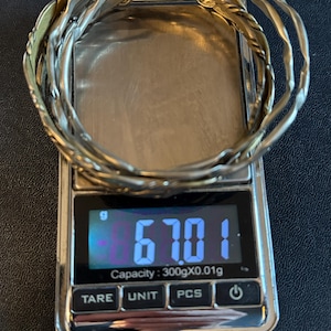 May include: A set of three metallic bangles with a twisted, textured design resting on a digital scale. The scale display shows a weight of 67.01 grams, with a maximum capacity of 300 grams and a precision of 0.01 grams clearly visible on the screen.