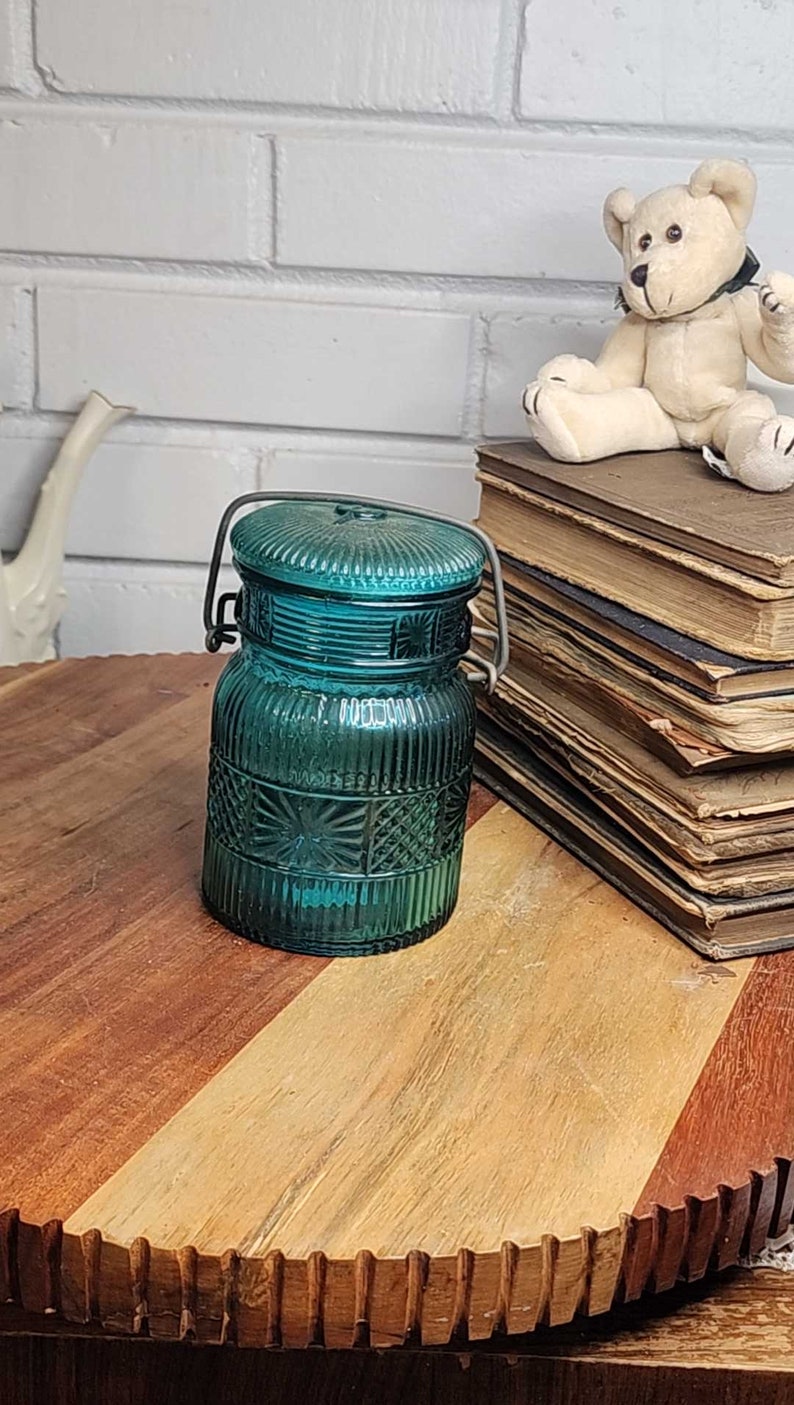 May include: A teal blue glass jar with a wire handle. The jar has a textured design and is sitting on a wooden surface.