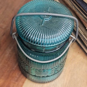 May include: A teal blue glass jar with a wire bail closure. The jar has a textured pattern and is sitting on a wooden surface.