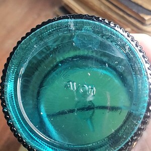 May include: Close-up of the bottom of a teal blue glass bottle with a textured rim. The bottle is marked with the word "Avon" in white lettering.