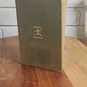 May include: A vintage hardcover book with a green cover and gold lettering. The book features a stylized figure on the cover. The book is displayed on a wooden surface.