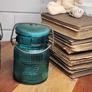 May include: Two teal blue glass jars with wire handles and a decorative pattern. The jars are sitting on a wooden surface with a stack of old books and a white teddy bear in the background.