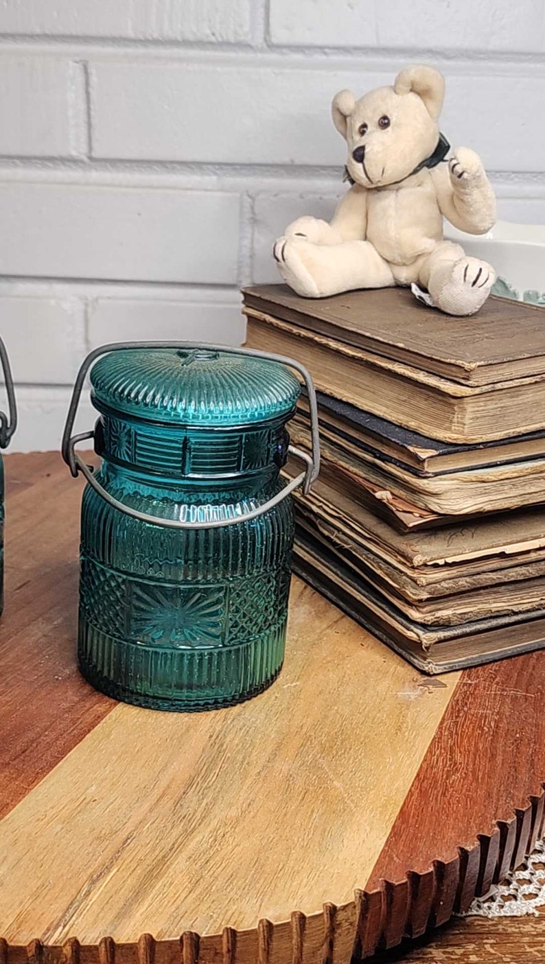 May include: Two teal blue glass jars with wire handles and a decorative pattern. The jars are sitting on a wooden surface with a stack of old books and a white teddy bear in the background.