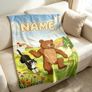 May include: A soft fleece blanket featuring a cartoon illustration of a brown bear, a black and white cat, a white chicken, and a white duck in a green meadow with colourful flowers under a blue sky. The word NAME is printed in large beige letters.
