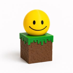 May include: A yellow spherical smiley face with black eyes and a curved mouth sits atop a brown cubic block with a green textured top layer. The brown base features a grid-like pattern of small squares, while the green top has a jagged edge.