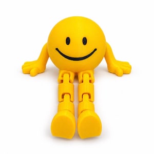 May include: A bright yellow, spherical figurine with a smiling face featuring black eyes and a black mouth. The figure has articulated arms extended to the sides and two jointed legs extending forward, all in a matching yellow color.
