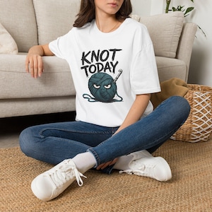May include: A white short-sleeved t-shirt featuring a graphic of a dark teal ball of yarn with an angry facial expression and a crochet hook. The text KNOT TODAY is printed in black above the yarn graphic in a bold, distressed font style.