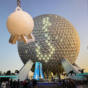 May include: A white, spherical keychain featuring a geometric, textured surface pattern and three support legs, suspended by a silver-toned metal chain. The object is positioned in front of a large, illuminated geodesic dome structure at dusk.