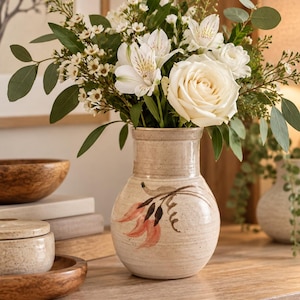 May include: A speckled beige ceramic vase with a narrow neck and a rounded body, featuring a hand-painted floral design in muted orange and dark brown. The vase is filled with a bouquet of white roses, alstroemeria, and green eucalyptus leaves.