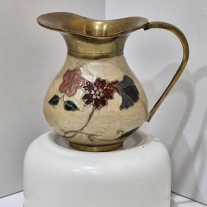 May include: A brass pitcher featuring a cream-coloured enamel body decorated with floral motifs in shades of red, pink, and dark blue. The pitcher has a curved handle, a wide pouring spout, and a polished brass rim and base, showcasing intricate metalwork.