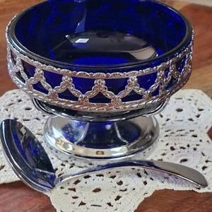 May include: A deep cobalt blue glass bowl set in a decorative silver-toned metal frame with a scalloped lattice pattern. The bowl rests on a circular silver-toned pedestal base and is accompanied by a matching silver-toned metal serving ladle.