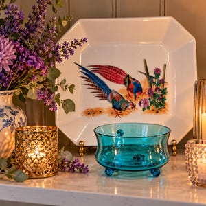 May include: A white octagonal ceramic platter featuring a colourful illustration of two pheasants among flowers, positioned behind a small, translucent teal glass bowl with three rounded feet and decorative glass spheres on its interior base.