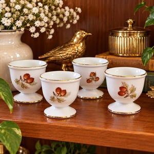 May include: A set of four white porcelain egg cups featuring a delicate botanical illustration of brown rose hips with green leaves. Each cup has a rounded bowl shape, a pedestal base, and a thin gold rim along the top edge and the base rim.