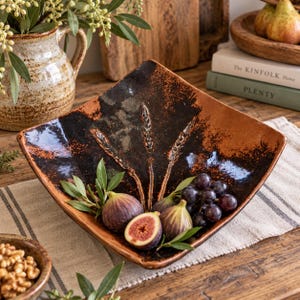 May include: A square, hand-crafted ceramic bowl with a dark brown and black glazed finish, featuring an embossed wheat stalk design. The bowl contains fresh purple figs and dark grapes, resting on a textured linen runner.