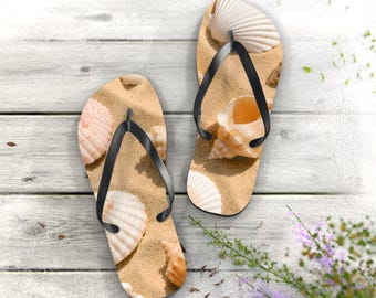 May include: A pair of flip-flops featuring a printed design of tan sand and various seashells in shades of white, cream, and light brown. The footwear has black rubber straps and a flat sole, showcasing a detailed beach-themed pattern.